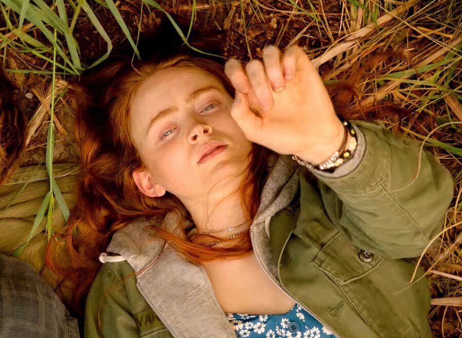 Sadie sink as mazzy lying in the grass while holding her hand up in a sacrifice